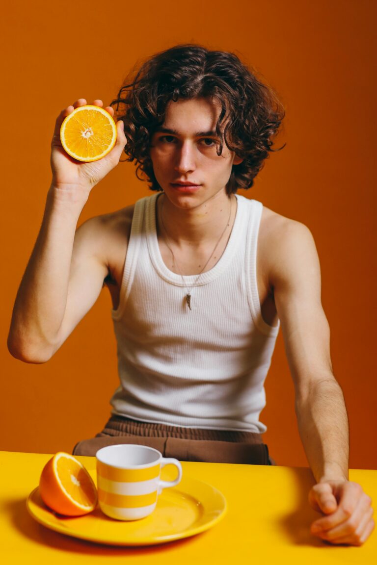 Stylish young man holding an orange slice against a vivid orange backdrop.